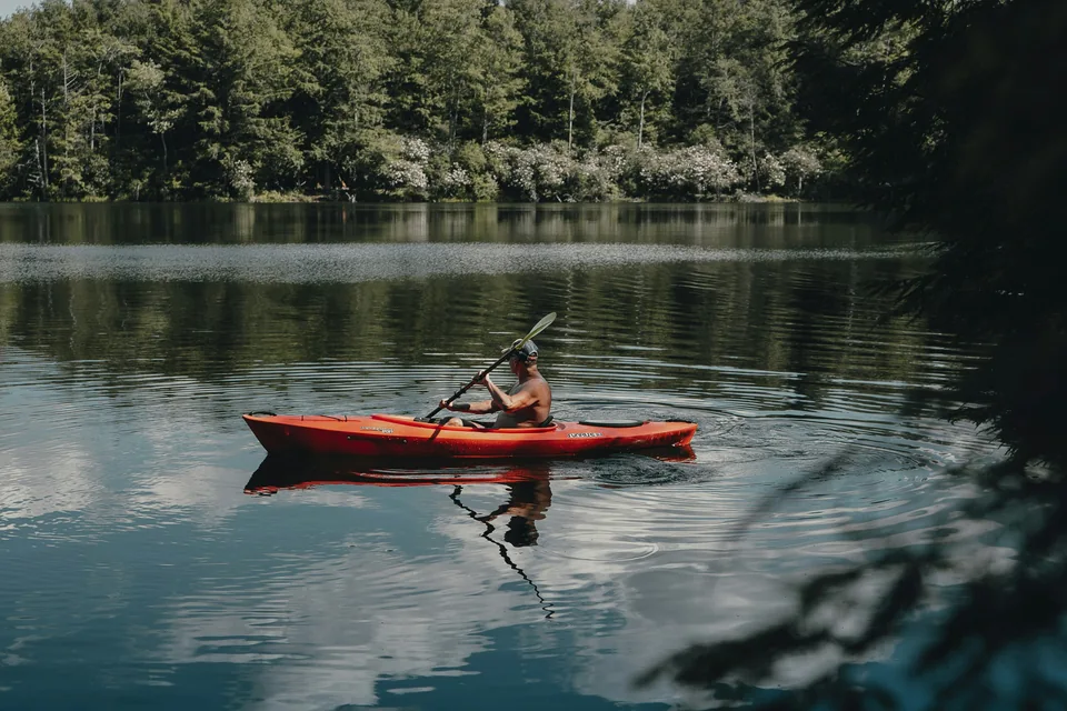 Un pratiquant pagaie dans un kayak rouge sur un lac bordé d’arbres.